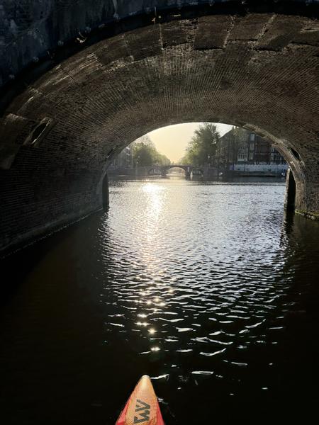 The photo captures a serene view from a kayak on the Amstel canal in Centrum, Amsterdam, looking through a historic brick arch bridge